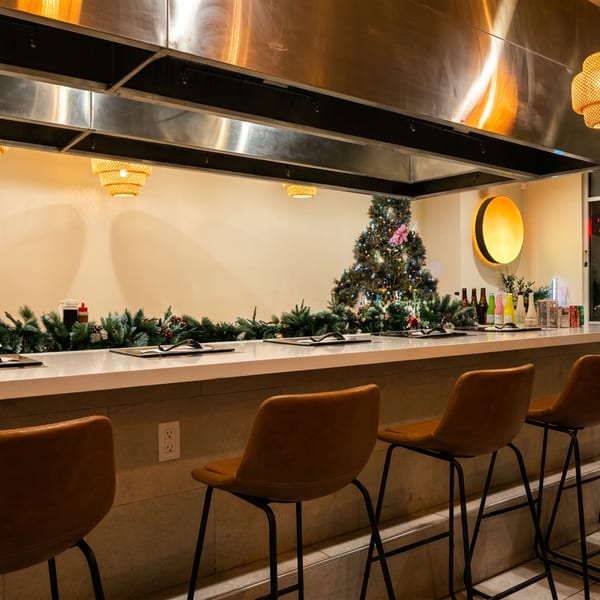 Modern restaurant counter with brown bar stools, festive Christmas tree, greenery, warm pendant lighting, and open kitchen hood.