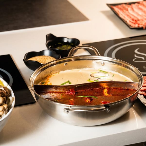 Divided hot pot with creamy and spicy broths, surrounded by sliced beef, vegetables, mushrooms, dipping sauces, and tofu.
