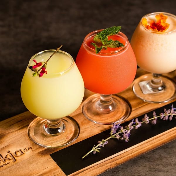 Three colorful cocktails garnished with herbs and flowers sit on a wooden serving board at a restaurant table.