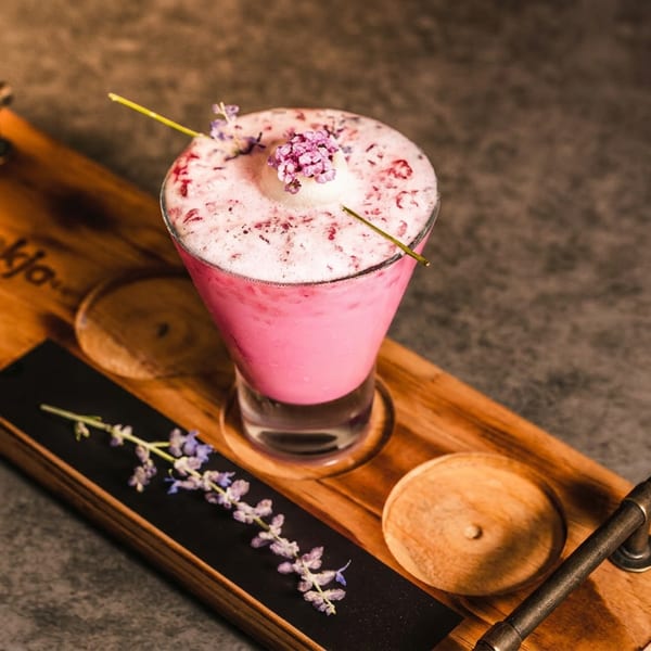 Pink frothy cocktail garnished with edible flowers, served on a wooden board with decorative floral sprigs.