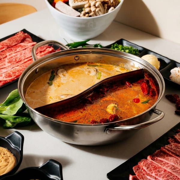 Split hot pot with spicy and creamy broths surrounded by marbled beef, rice, vegetables, mushrooms, and dipping sauces.