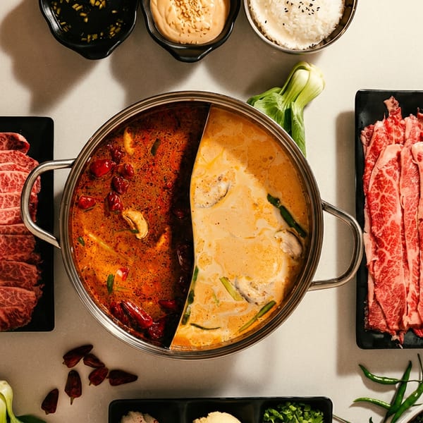 Overhead spread featuring a divided hot pot with spicy and creamy broths, marbled beef slices, sauces, rice, and vegetables.
