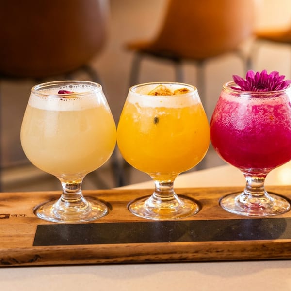 Three colorful beverages in stemmed glasses sit on a wooden serving tray at a warmly lit restaurant table.