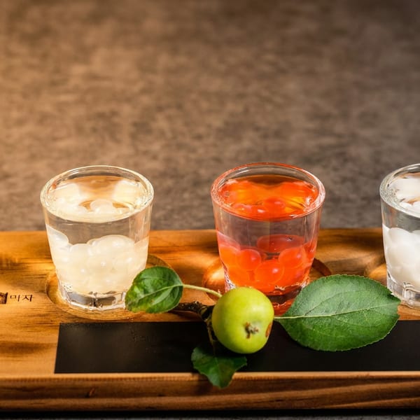 Three colorful fruit drinks with ice sit on a wooden tray beside green leaves, branded Moojigae Shabu Shabu Restaurant.